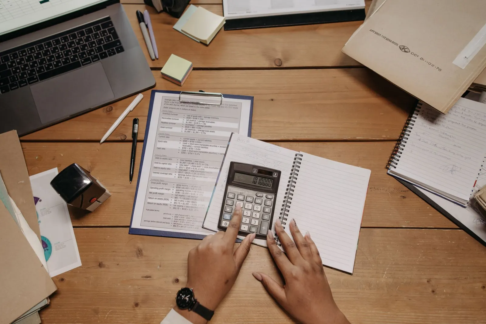 Des mains utilisent une calculatrice près de documents et d’un ordinateur portable sur un bureau en bois.